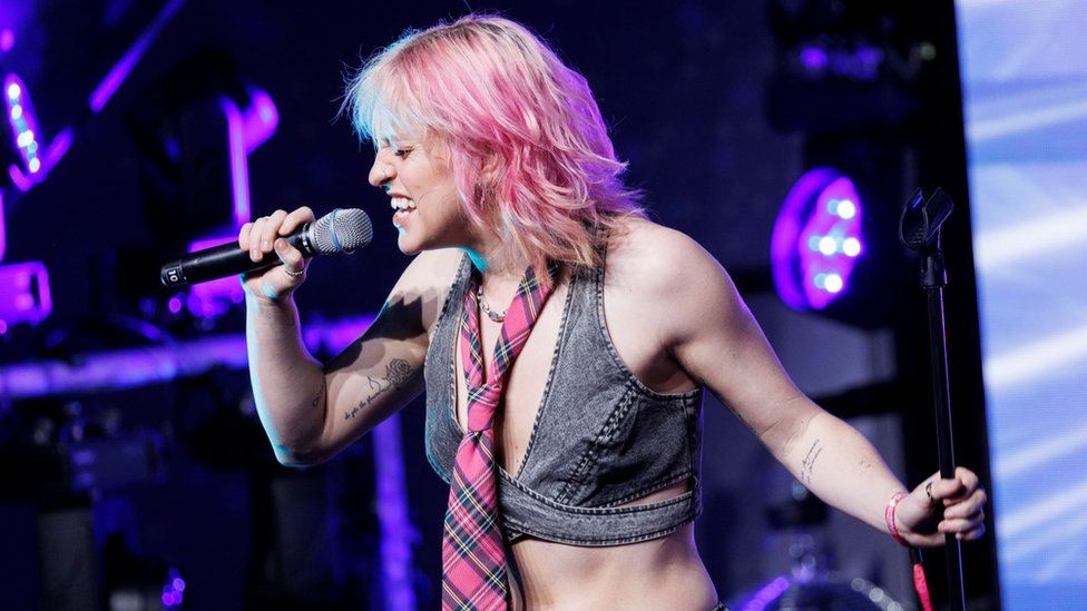 Beth McCarthy performing on stage. Beth is a 26-year-old white woman with blonde hair dyed pink. She wears a grey denim crop top with a pink tartan tie around her neck. She holds a microphone to her face with her right hand, revealing a black line tattoo on her inner upper arm. Beth's eyes are closed as she sings, holding the microphone stand with her left hand. The staging behind her is lit purple