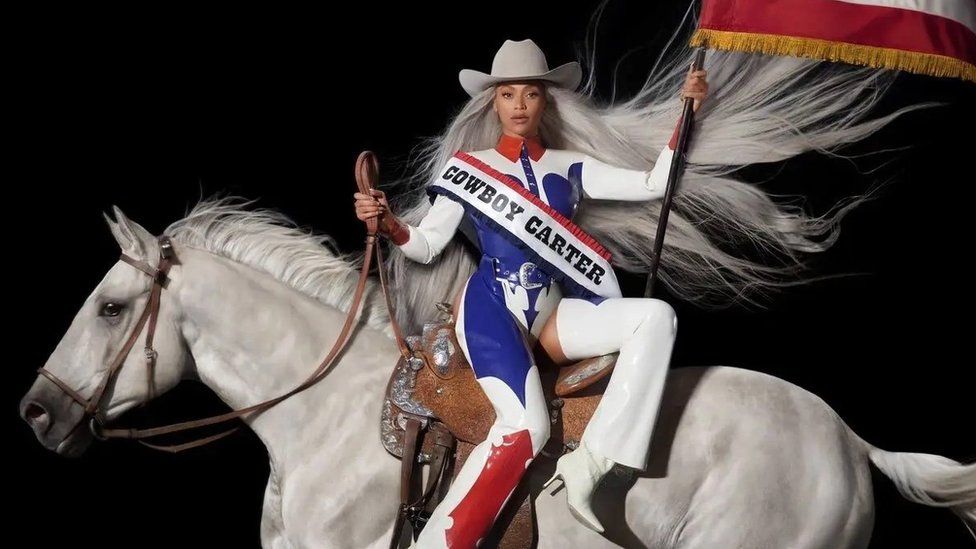 Album art for Beyoncé's album, Cowboy Carter. Beyoncé is a 42-year-old black woman. She's styled in a red, white and blue patent cowboy-themed outfit including a cowboy hat, chaps and a buttoned up shirt. She's pictured sitting side-saddle on a white horse, her long silver hair flowing behind her. In her right hand, she holds the horse's reigns and in her left she holds aloft the American flag.